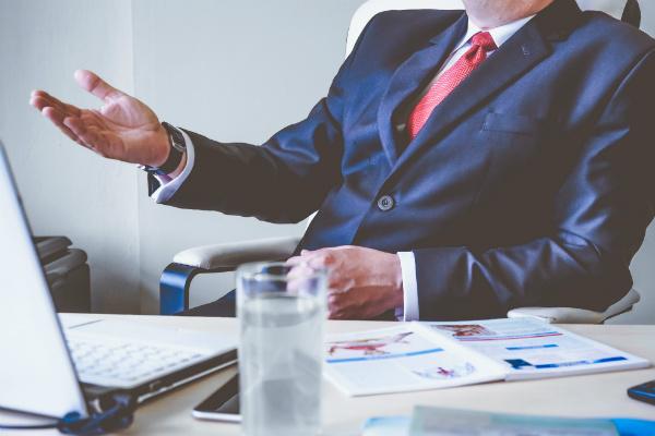 man in a blue suit at his desk, gesturing with his hands